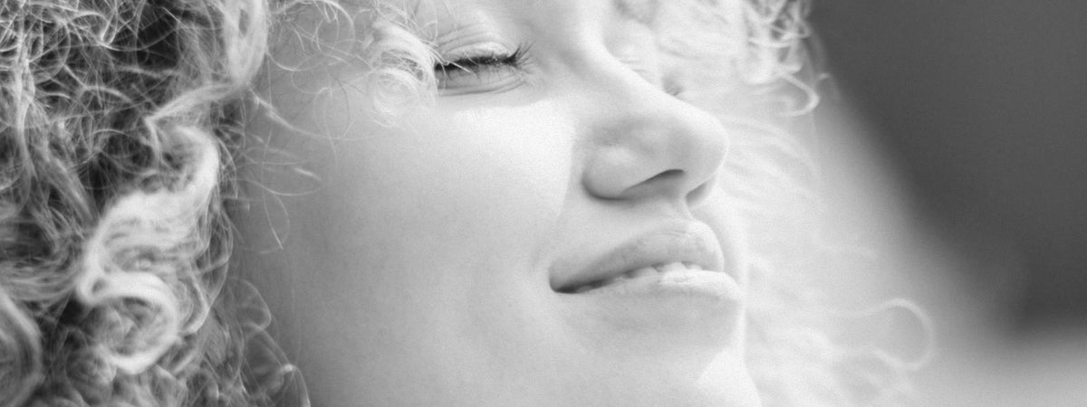 A person with curly hair smiling with their eyes closed, face turned to the light, black and white